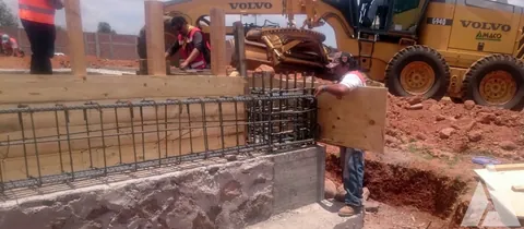 Trabajadores armando el refuerzo y la cimbra de madera de un muro con maquinaria Volvo detrás