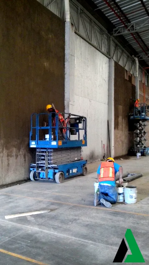 Trabajadores con plataformas de tijera aplicando acabado al muro interior de una nave industrial con cubierta metálica