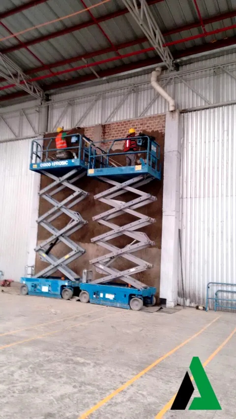 Plataformas de tijera frente a un muro de ladrillo en preparación dentro de una nave industrial con estructura metálica