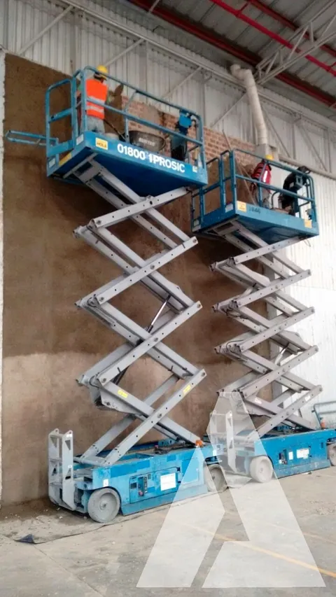 Dos plataformas de tijera elevadas con trabajadores acabando el muro interior de una nave industrial de gran altura