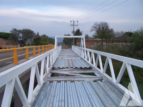 Puente peatonal de estructura metálica blanca con tablero de lámina losacero recién montado junto a un puente vehicular