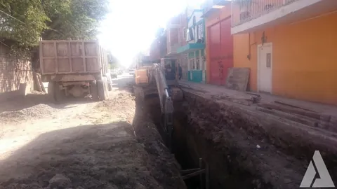 Zanja profunda para colector de alcantarillado excavada a lo largo de una calle con casas de colores, con camión de volteo y excavadora